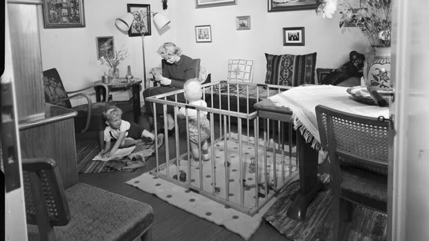 Husmor med barn i hemmiljö. Trångt vardagsrum med en kvinna, två barn och en barnhage. 1940-tal.