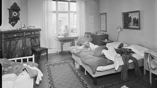 Husmor med barn i hemmiljö. Ett barn sitter i fönstret i ett sovrum med obäddade sängar. 1940-tal